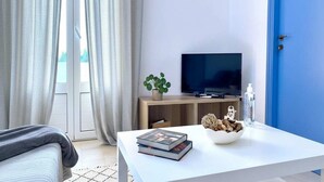 Una habitación moderna con una televisión sobre un soporte de madera, una mesa blanca con libros y un cuenco decorativo, una puerta azul y una ventana con cortinas.