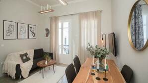 Ett modernt vardagsrum med en soffa, ett soffbord och ett matbord. På väggen hänger inramade konstverk, det finns en stor spegel och ett fönster med gardiner.