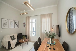 A modern living room with a sofa, coffee table, and dining table. There are framed artworks on the wall, a large mirror, and a window with curtains.