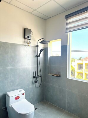 A bathroom with a toilet, a shower with a handheld showerhead, a wall-mounted control panel, and a window with a view of buildings. - The Sea Homestay Phan Thiet (Phan Thiet)