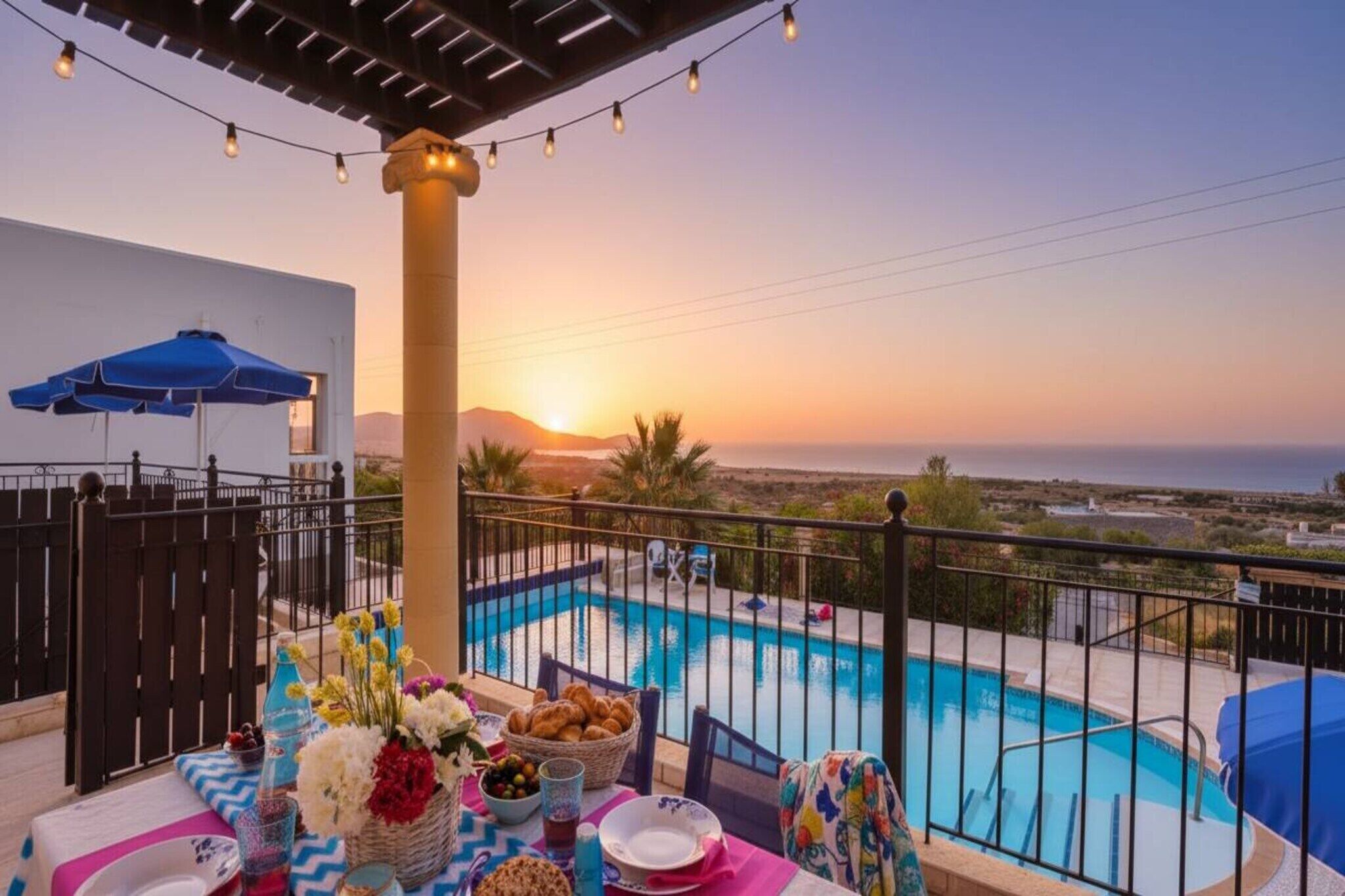 Escena junto a la piscina con vistas al atardecer, una mesa con comida y bebidas, y una pérgola con luces colgantes.