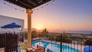 A poolside setting with a sunset view, a table with food and drinks, and a pergola with string lights. - Pool Villa Overlooking the Sea (kalathos)