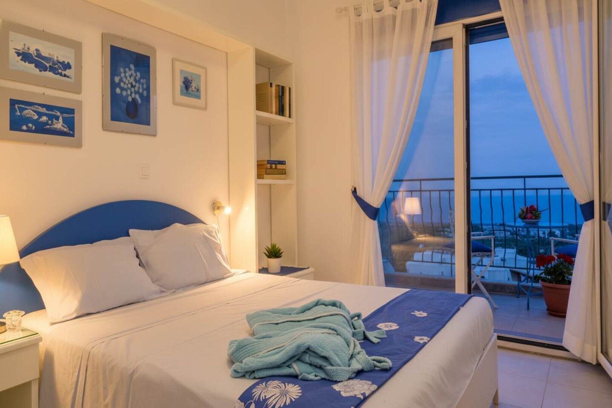 Un dormitorio con cama, mesita de noche, cortinas en la ventana y un balcón con una silla, con vista al mar.