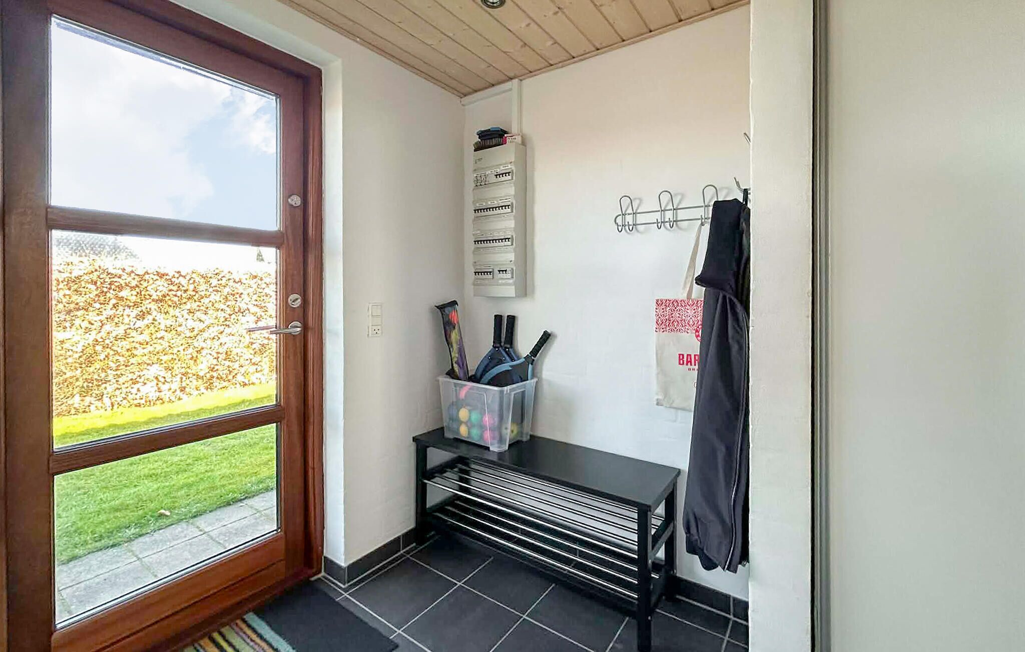 Een kamer met een houten deur, een betegelde vloer, een bankje, een wandrek en uitzicht op een grasveld.
