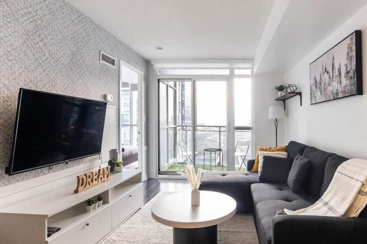 A modern living room with a large flat-screen TV, a sofa, a coffee table, and a balcony with a view of the city.