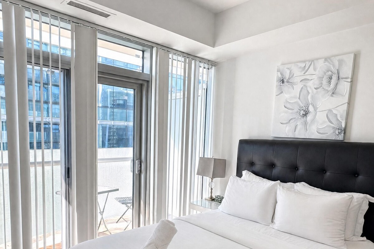 A hotel room with a large bed, a headboard, a window with blinds, a painting on the wall, and a view of the outside.
