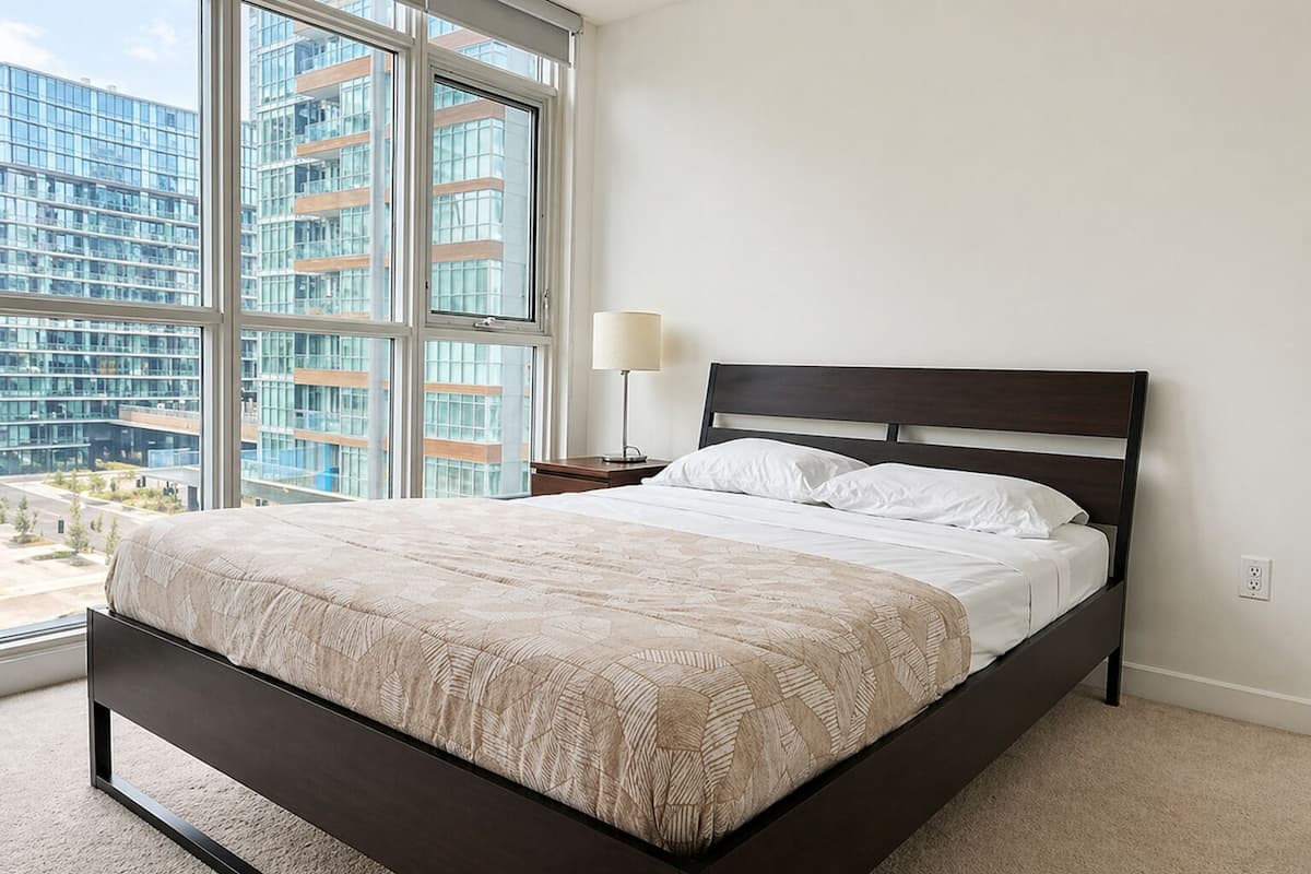 A modern bedroom with a large bed, a nightstand, a lamp, and a view of a cityscape through large windows.