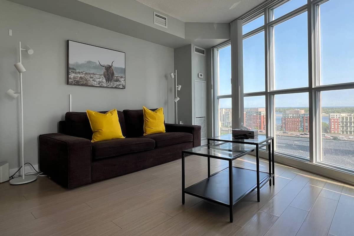 A modern living room with a dark brown sofa, two yellow cushions, a glass coffee table, and a large window offering a city view.