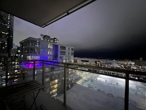 Un immeuble de grande hauteur doté d’une façade en verre, d’un balcon avec une piscine illuminée de lumière bleue, et offrant une vue sur le paysage urbain la nuit.