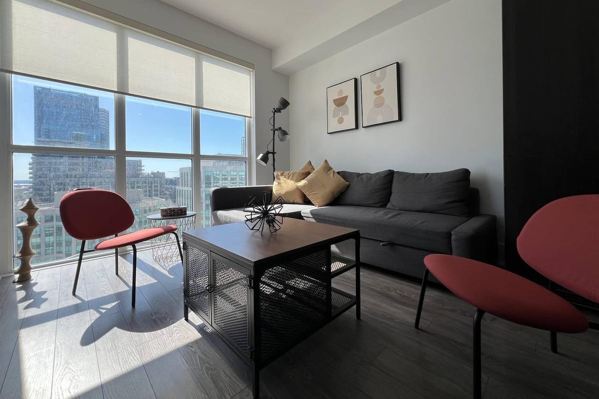 A modern living room with a grey sofa, red chairs, a coffee table, and a large window offering a city view.