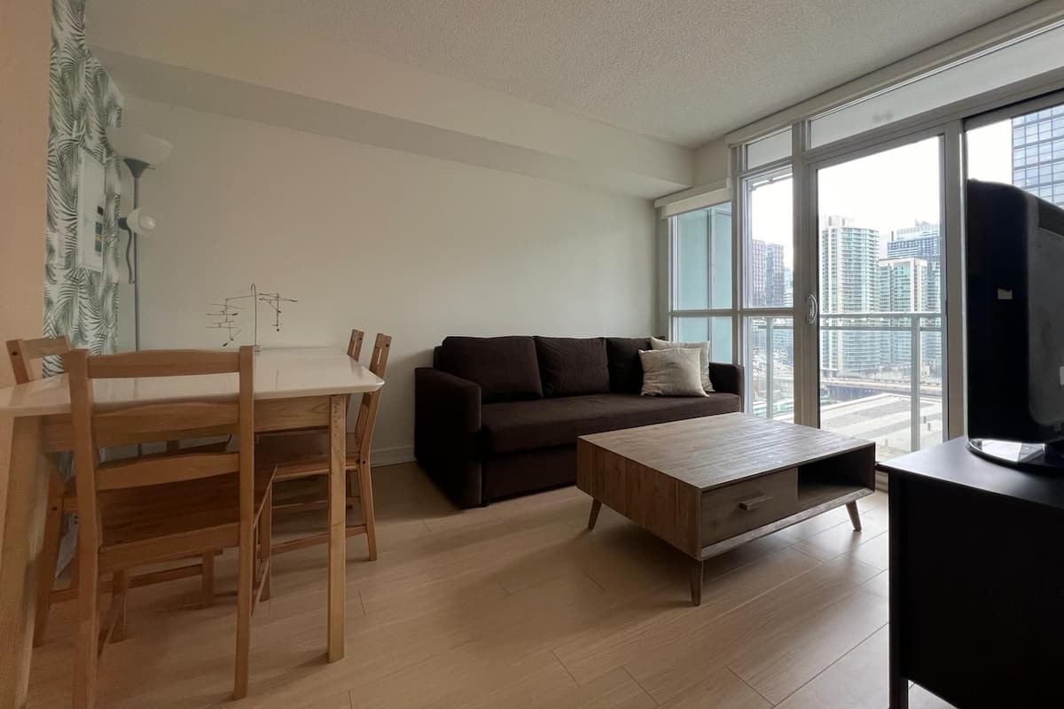 A modern living room with a dining table, wooden chairs, a brown sofa, a wooden coffee table, and a large window offering a city view.