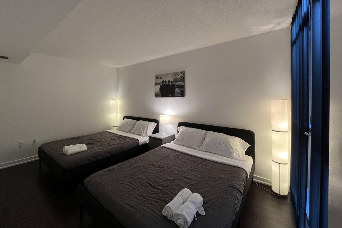 A hotel room with two beds, each with a pillow and a rolled towel. There is a framed picture on the wall and a lamp on each side of the beds.