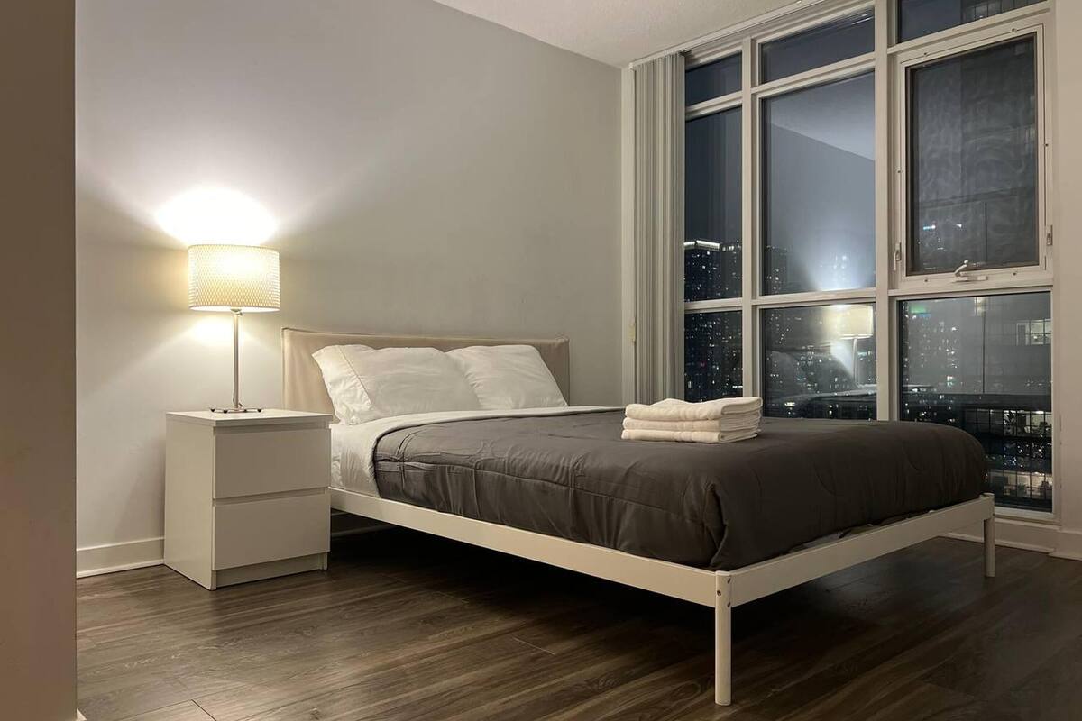 A bedroom with a bed, a nightstand, a lamp, and a large window with a city view.