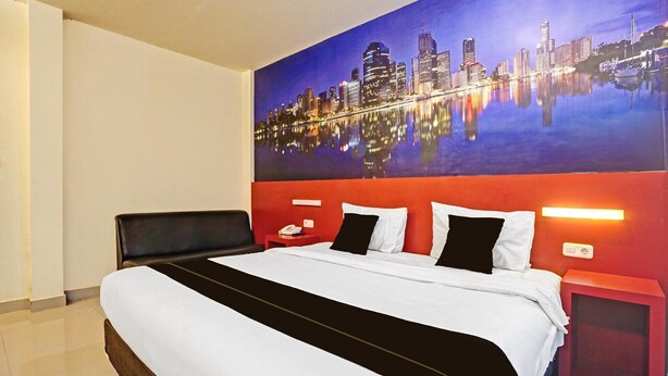 A hotel room with a large bed, a cityscape mural on the wall, a red accent wall, and a nightstand with a phone.