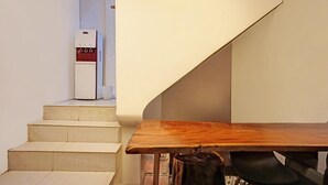 Un escalier menant à une pièce où se trouvent une table en bois et une chaise, un petit appareil électroménager blanc sur le palier, et un sac noir sur le sol.