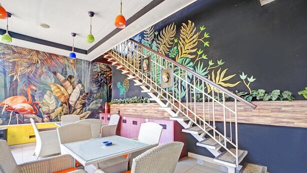 Ein Restaurant mit Korbmöbeln, einer Treppe mit Geländer sowie einer Wandmalerei mit tropischen Blättern und Flamingos.