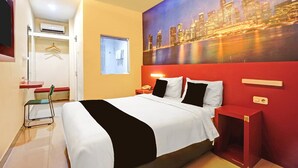 Habitación de hotel con cama, pintura de paisaje urbano, pared de acento roja, mesita de noche pequeña y una silla.