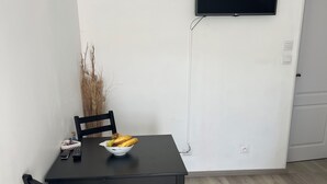A dining area with a black table, two chairs, a bowl of fruit, a remote control, and a wall-mounted TV.