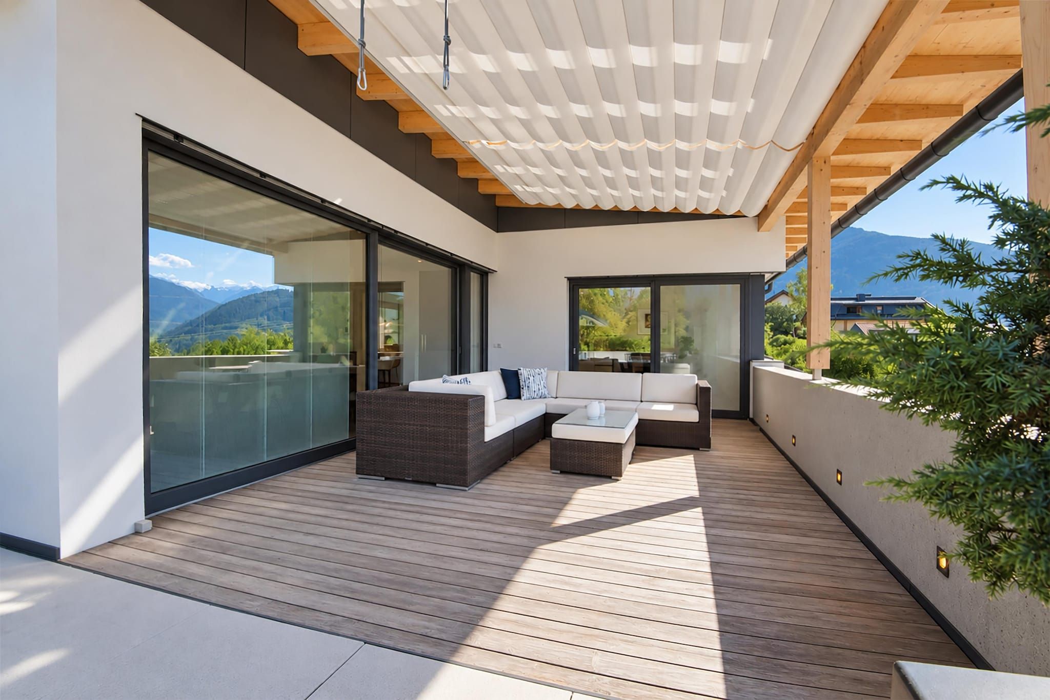 Un espace de vie extérieur moderne, doté d’une terrasse en bois, de sièges intégrés et de grandes portes-fenêtres coulissantes offrant une vue sur les montagnes.