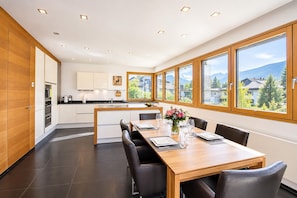 A modern kitchen with white cabinets, a wooden dining table, and a view of houses through large windows.