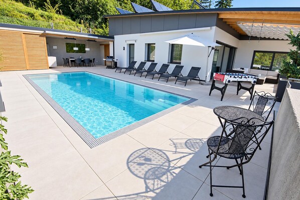 Un espace piscine extérieur moderne, doté d’une piscine à l’eau cristalline, de chaises longues, d’une table de baby-foot et d’un coin salon abrité.