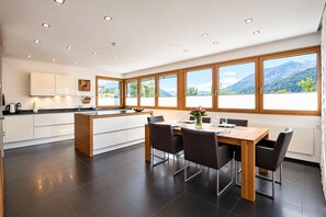 Una cocina moderna con una isla central, un comedor con mesa y sillas, y grandes ventanales con vistas a la montaña.