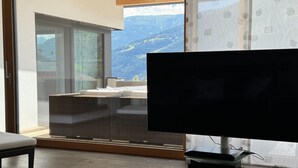 A modern living room with a large flat-screen TV, a sofa, and a glass partition leading to a balcony with a mountain view. - Villa in Tyrol with pool (Stanz bei Landeck)