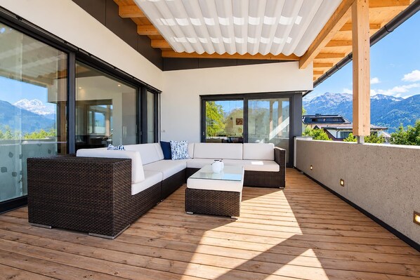 Un área exterior moderna con terraza de madera, un conjunto de sofás y una pared de vidrio con vista a la montaña.