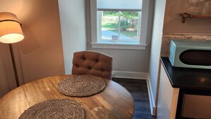 Een eetgedeelte met een ronde tafel, twee geweven placemats, een gestoffeerde stoel, een lamp, een raam met uitzicht en een magnetron.