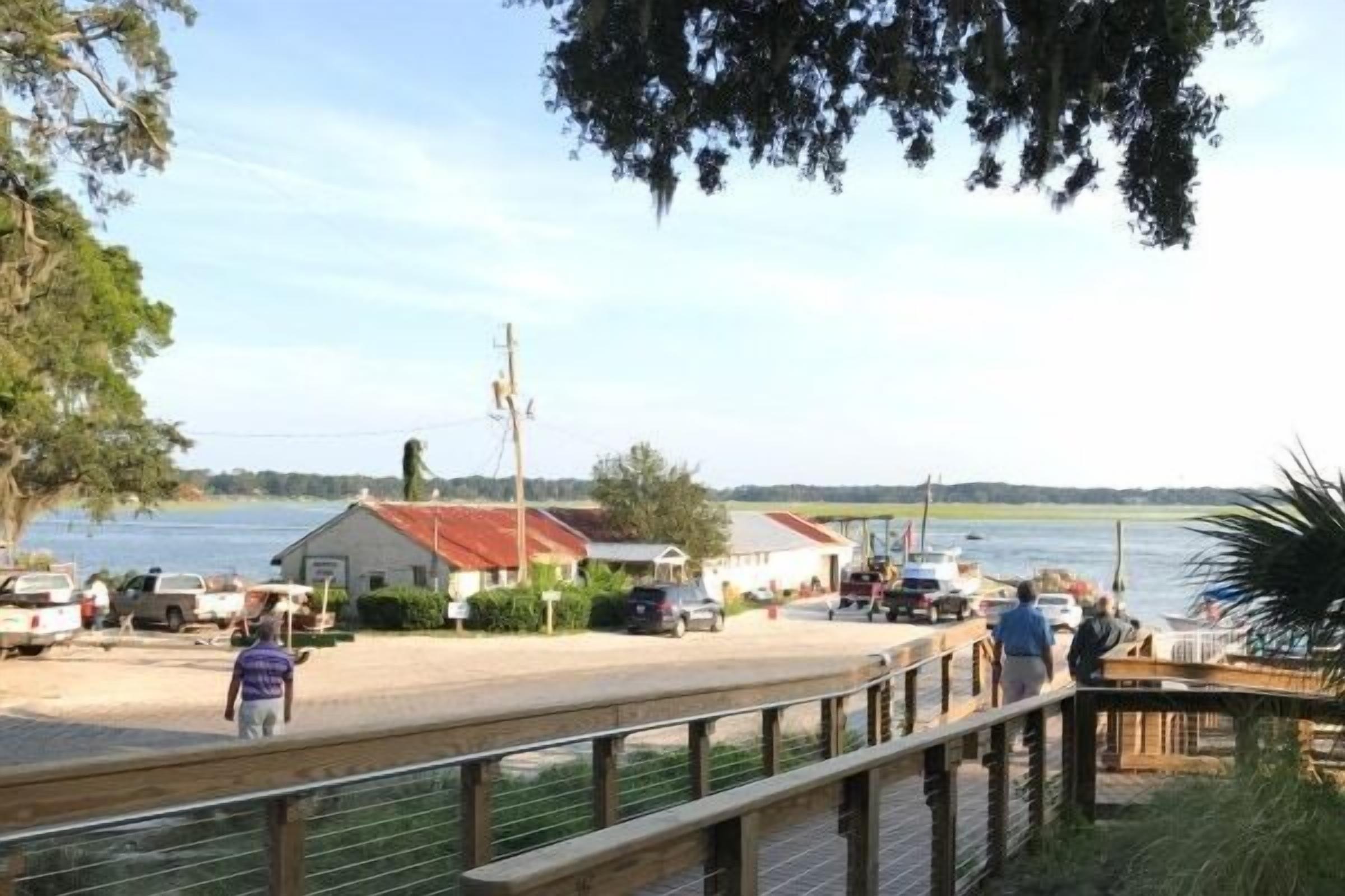 A waterfront property with a dock, a small building with a red roof, and a paved area with vehicles.