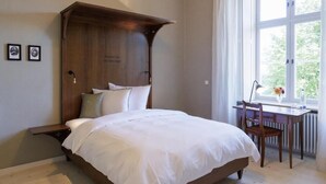 A bedroom with a wooden headboard, a bed with white linens, a desk with a chair, and a window with a view of greenery. - Kloster Beuerberg  (Beuerberg)