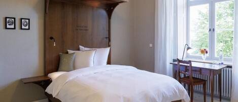 Un dormitorio con cabecera de madera, cama con ropa de cama blanca, escritorio con silla y ventana con vista a la vegetación.