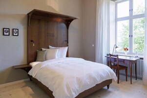 A bedroom with a wooden headboard, a bed with white linens, a desk with a chair, and a window with a view of greenery.