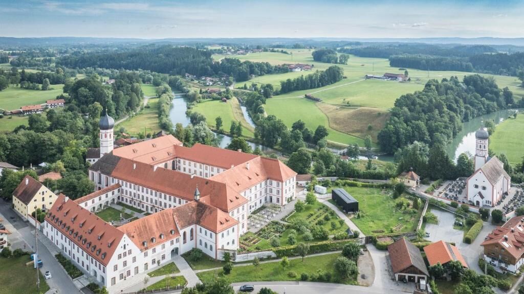 An aerial view of a large, historic building complex with multiple wings, surrounded by lush green fields and a river.