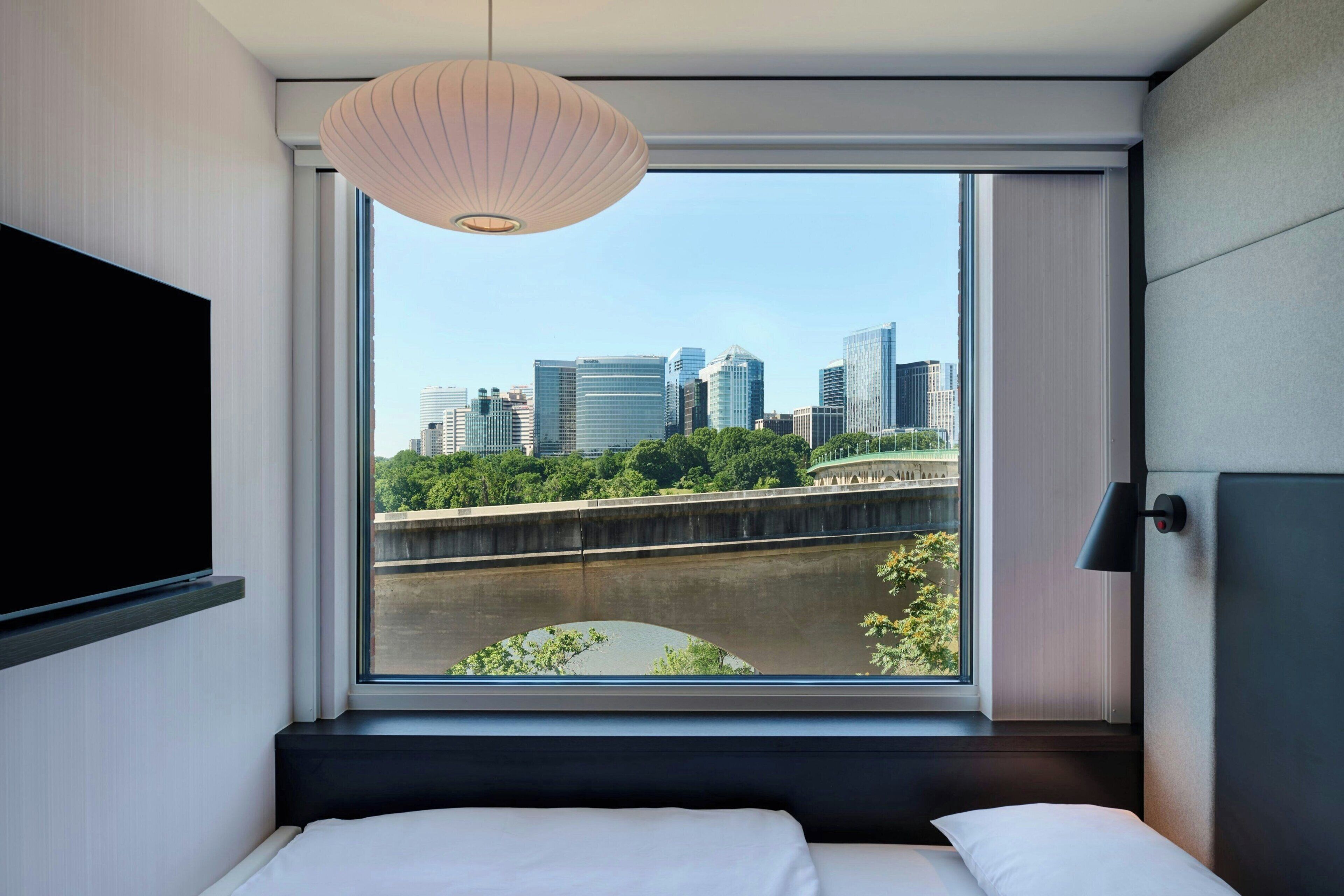 A hotel room with a large window, a bed, a TV, and a hanging light fixture.
