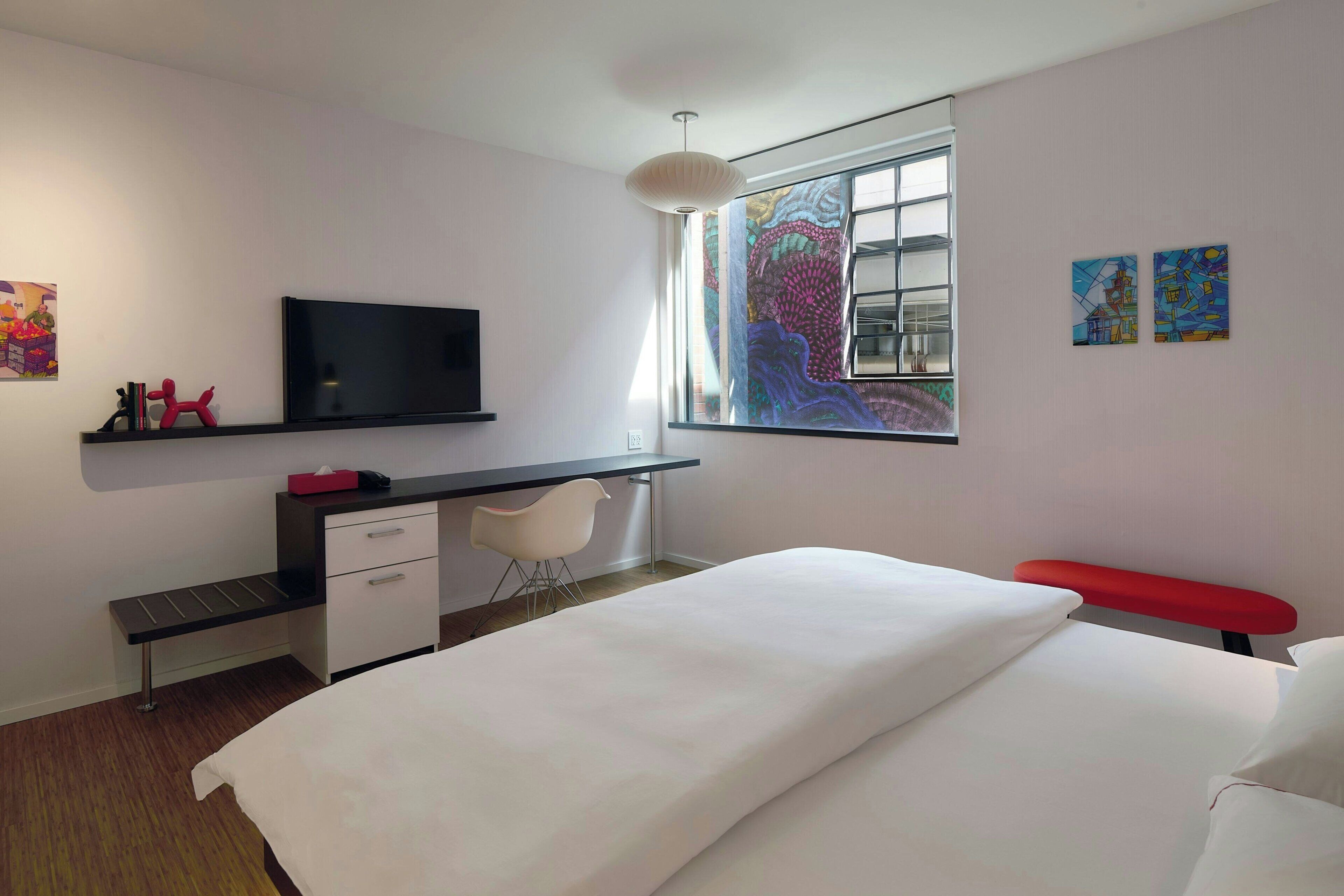 A modern bedroom with a bed, a desk, a chair, a television, and artwork on the walls.