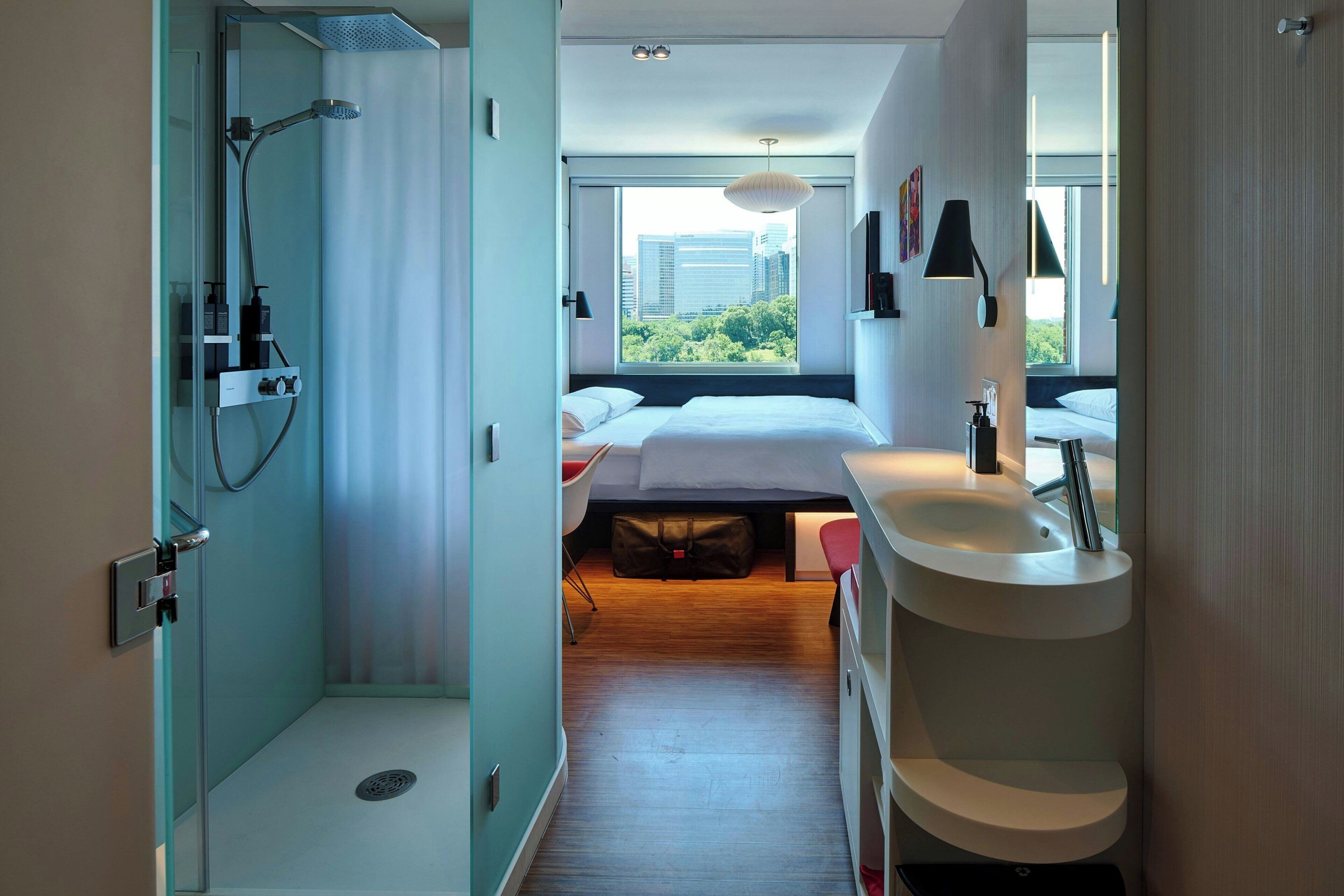A modern hotel room with a shower, a bed, a desk, and a view of the city.