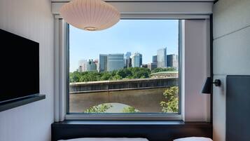 A hotel room with a large window, a bed, a mounted TV, and a hanging light fixture.
