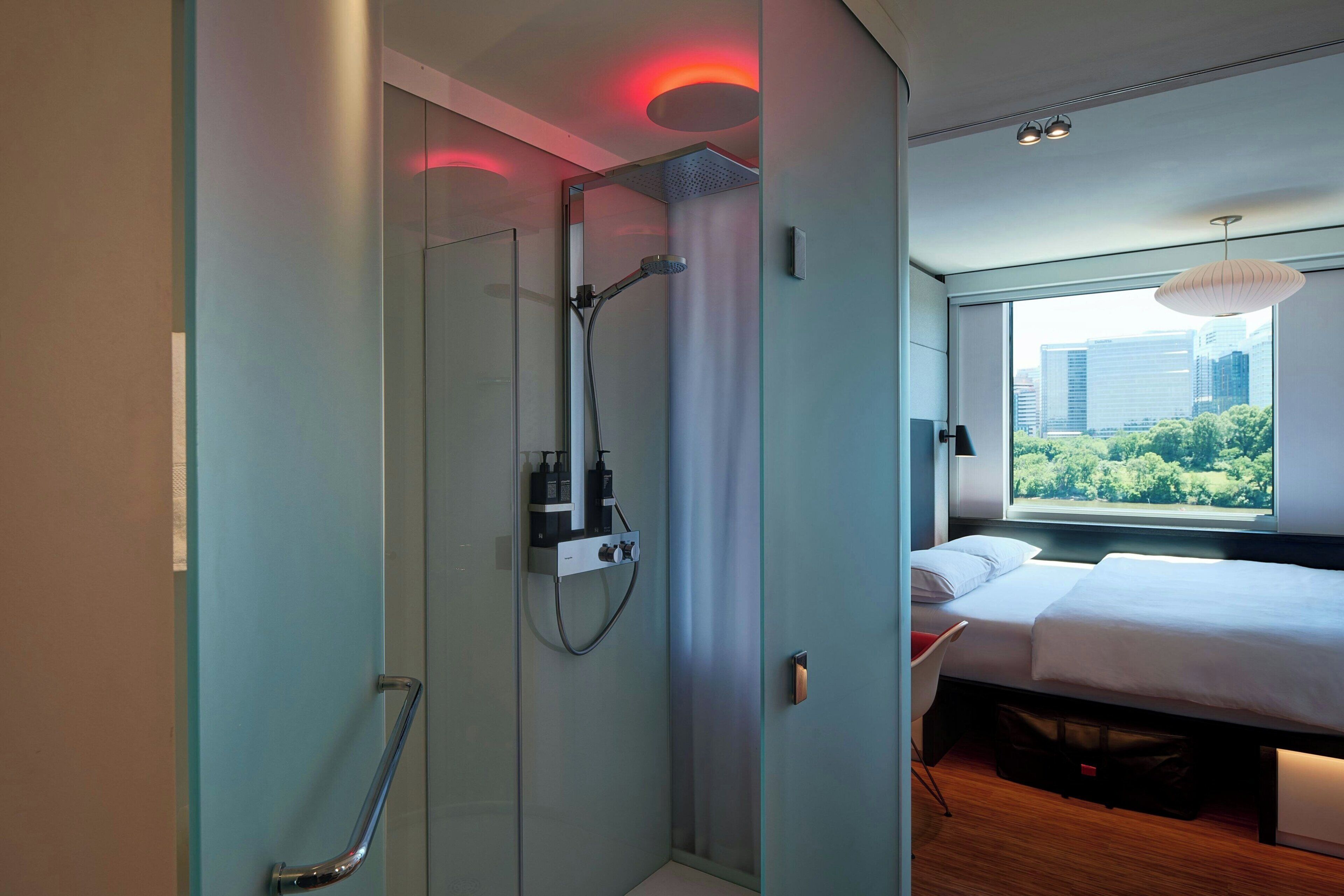 Ein modernes Hotelzimmer mit einer Duschkabine aus Glas, einem Bett mit weißer Bettwäsche und Blick auf die Stadt durch ein großes Fenster.