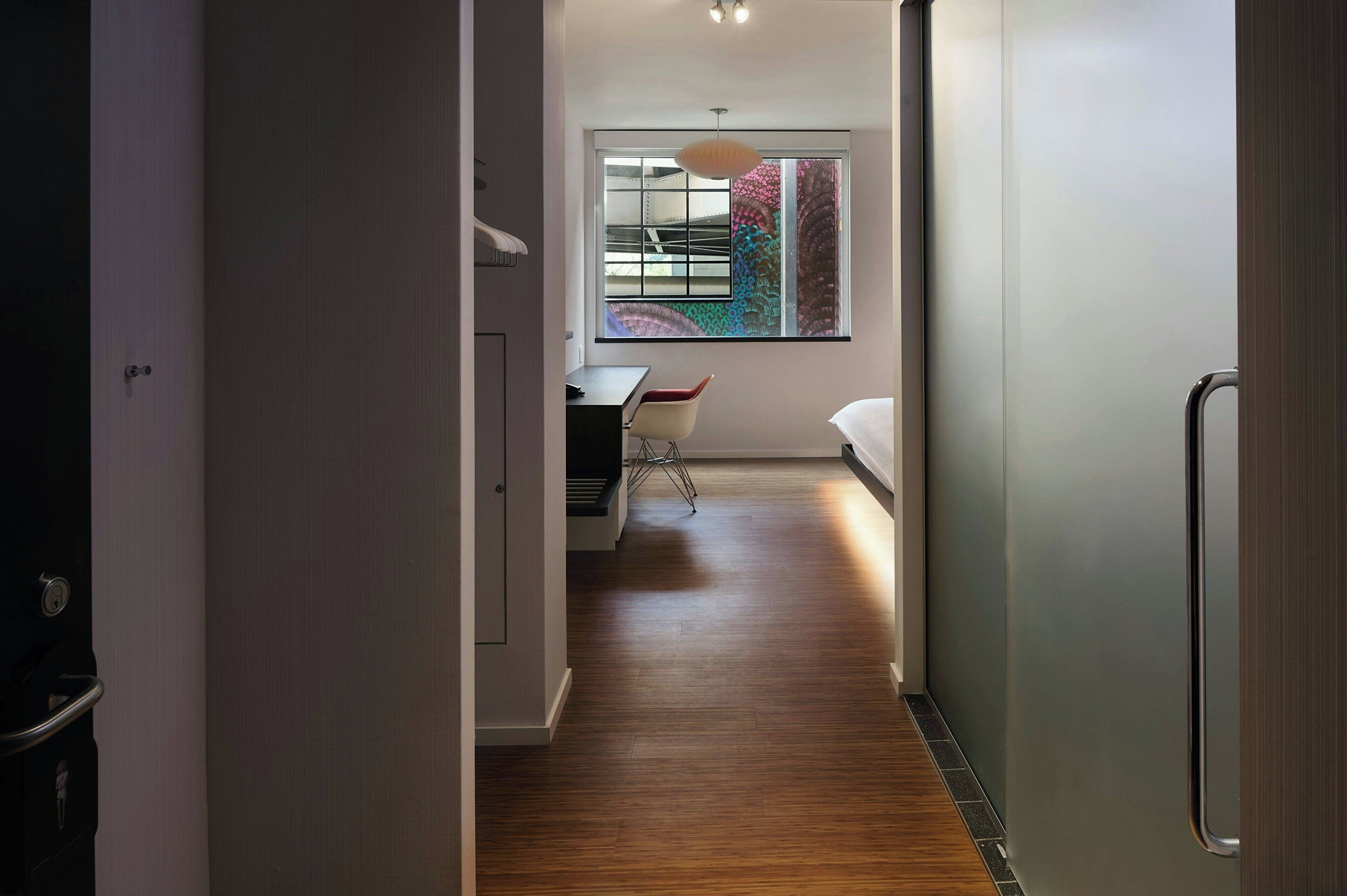 Un couloir mène à une chambre dotée d’un plancher en bois, d’un bureau avec une chaise et d’un lit.