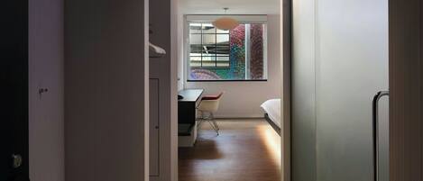 Un couloir mène à une chambre dotée d’un plancher en bois, d’un bureau avec une chaise et d’un lit.