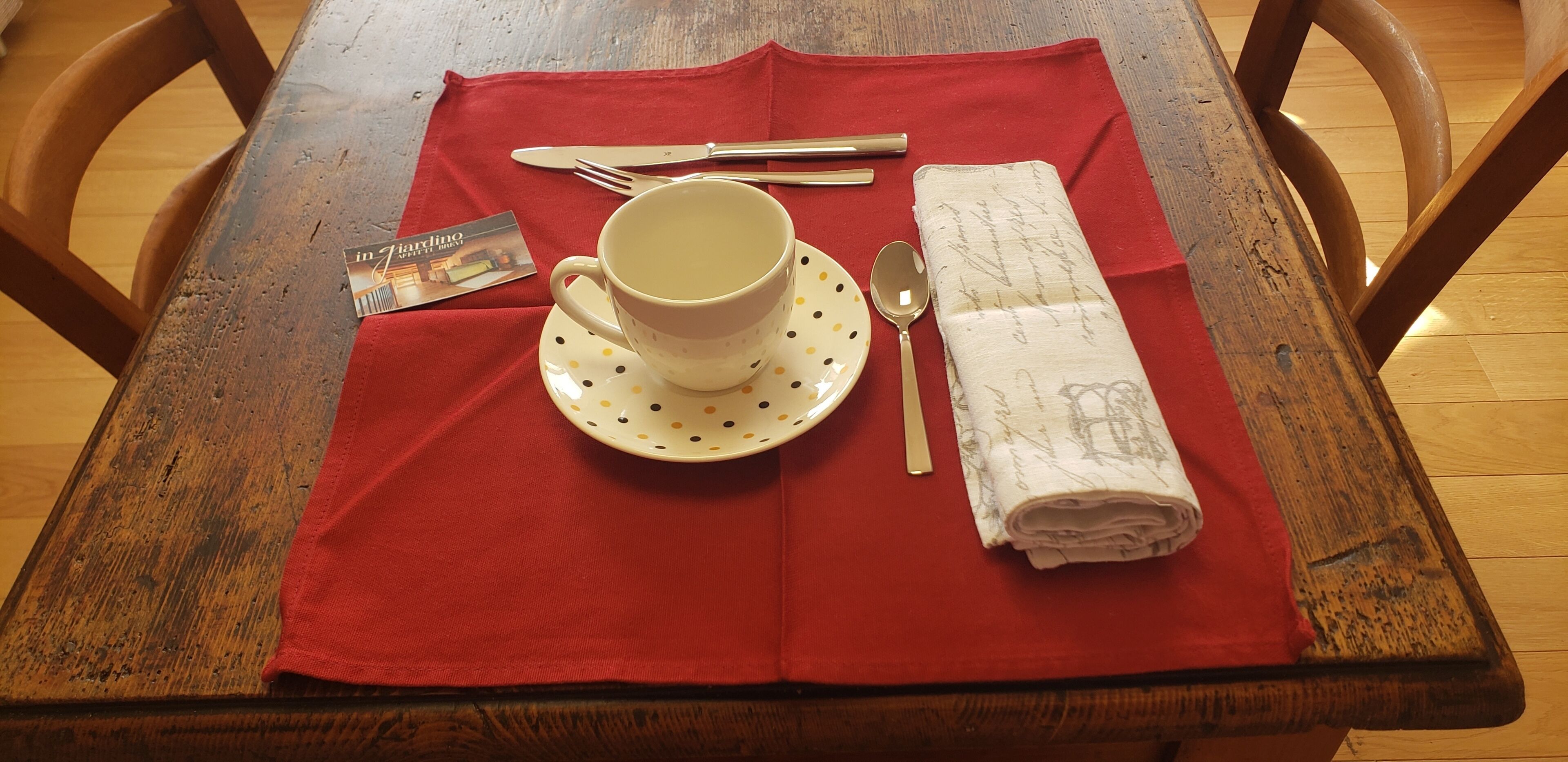 Una mesa de madera con un mantel rojo, una taza blanca sobre un platillo, cubiertos, una cuchara, una servilleta doblada y una revista.