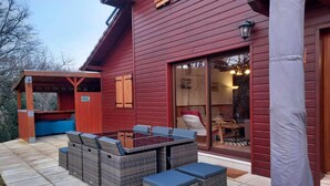 A red wooden building with a glass door entrance, a covered patio with a table and chairs, and a stone wall. - Cottage in Lachapelle-Auzac with Pool (LACHAPELLE AUZAC)