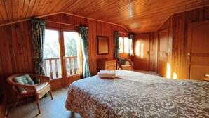 A wooden cabin bedroom with a bed, a chair, a dresser, and a window with curtains. - Cottage in Lachapelle-Auzac with Pool (LACHAPELLE AUZAC)