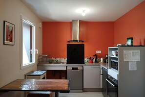 A compact kitchen with a stove, oven, and dishwasher, a window with blinds, and a wooden table. - Modern Tiny House in Souillac (SOUILLAC)
