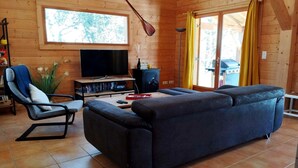 A wooden cabin interior with a sofa, a chair, a television, a fireplace, and a window with curtains. - Home with Enclosed Garden near Sarlat (PECHS DE L'ESPERANCE)
