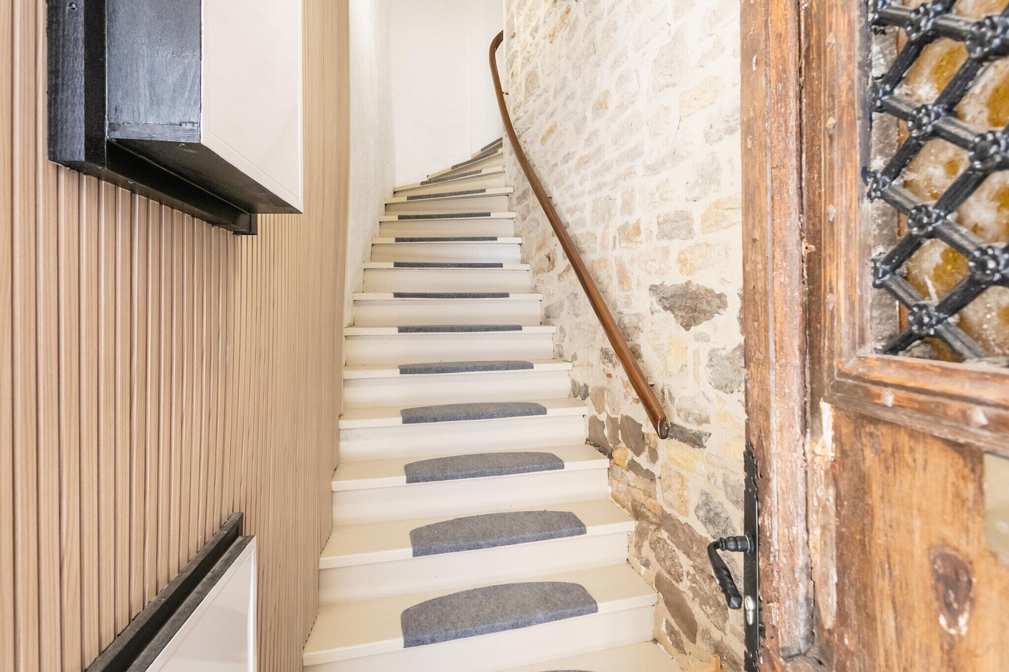 Un escalier en colimaçon avec des marches en pierre et une rampe en bois, menant à une pièce dotée d’un mur en pierre et d’une porte en bois ornée d’un motif en treillis.