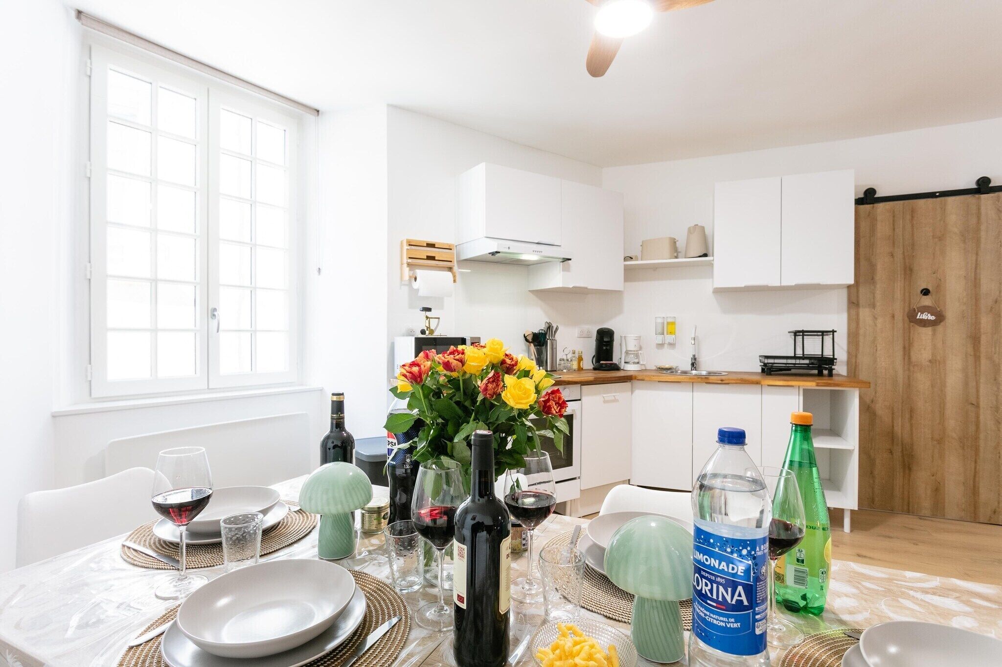 A dining area with a table set for two, a vase of flowers, and various bottles.