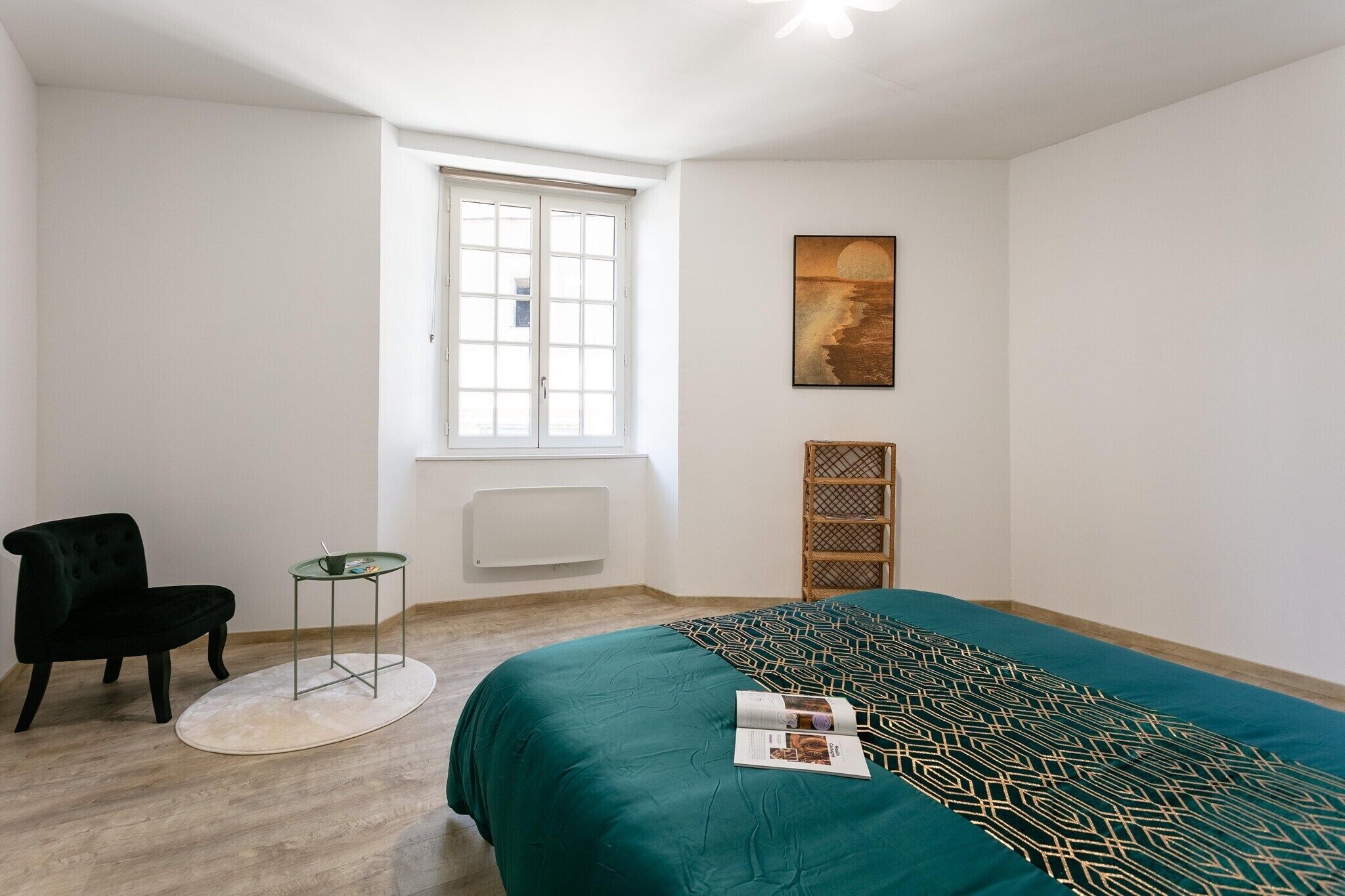 Ein Schlafzimmer mit einem Bett, einem Sessel, einem kleinen Tisch, einem Fenster und einem Bild an der Wand.