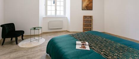 Ein Schlafzimmer mit einem Bett, einem Sessel, einem kleinen Tisch, einem Fenster und einem Bild an der Wand.